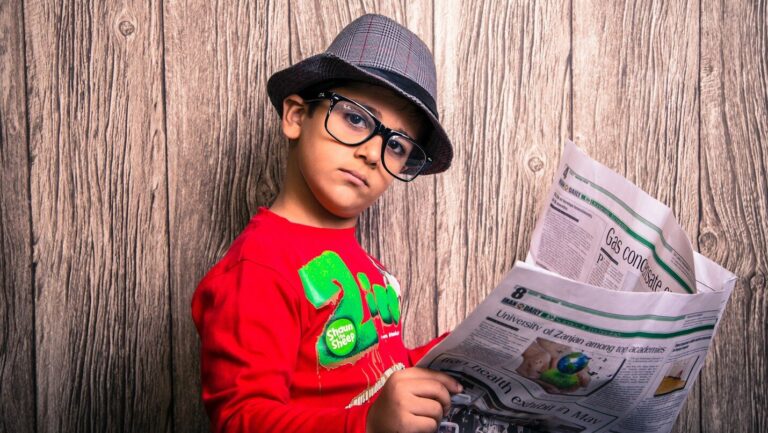 a young boy wearing glasses and a hat is reading a newspaper and looking at the camera skeptically